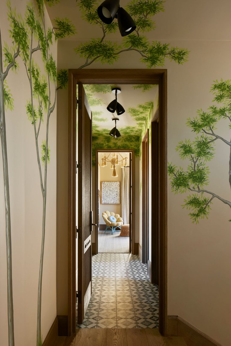 A hallway with painted walls and ceiling features realistic green tree branches. Decorative tile flooring runs through the corridor, leading to a sunlit seating area with blue chairs and gold chandeliers. Black ceiling light fixtures line the hallway.