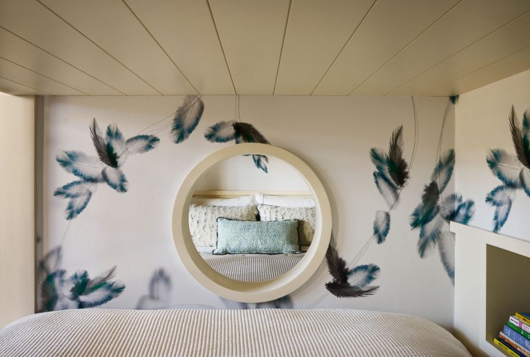 A cozy bedroom with a circular mirror above the bed, reflecting a pillow and headboard. The wall is decorated with a feather pattern in blue, black, and white, creating a serene atmosphere. Part of a bookshelf with books is visible on the right side.