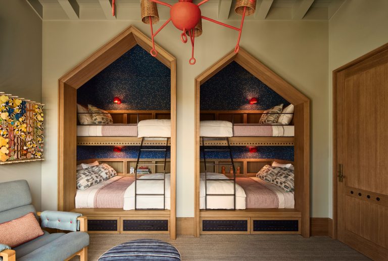 A cozy bedroom features two built-in bunk beds set in arched wooden alcoves with navy starry wallpaper, each bed with white bedding and patterned pillows. A playful red ceiling light hangs above, and a chair and ottoman sit in the corner.