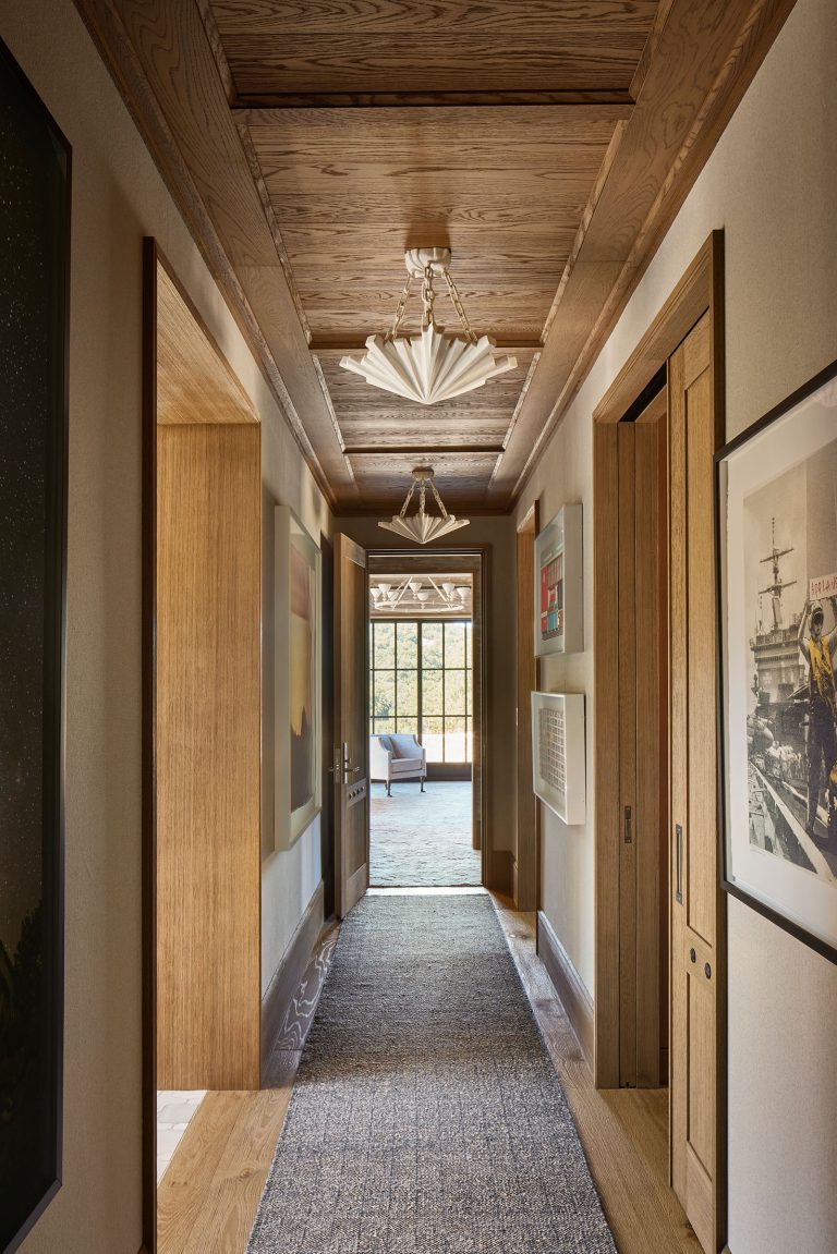 A long hallway with wood-paneled ceiling and doors, beige walls, and framed art on both sides. A textured gray runner covers the wooden floor. At the end, sunlight streams through a glass door, revealing greenery and an outdoor seating area.