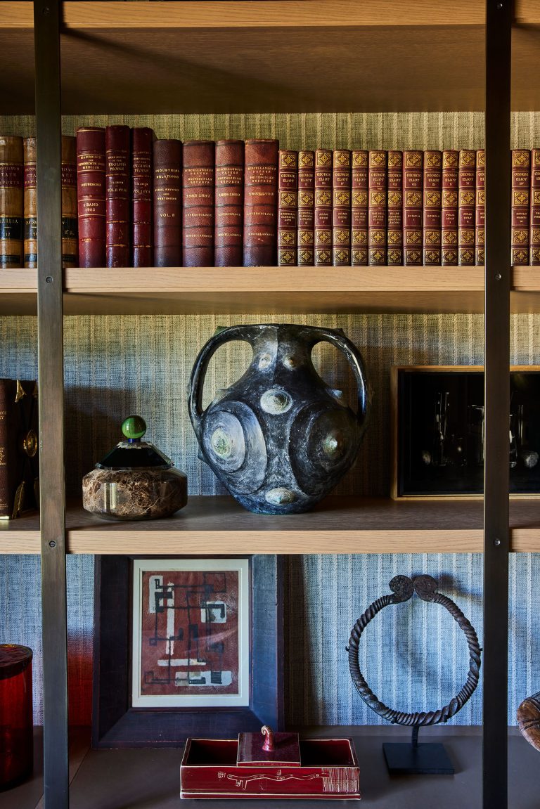 A bookshelf with rows of old leather-bound books, a dark metallic sculptural vase, a decorative round box, framed abstract art, a twisted metal bracelet, and a small red box, all arranged on wooden shelves with a textured wallpaper backdrop.