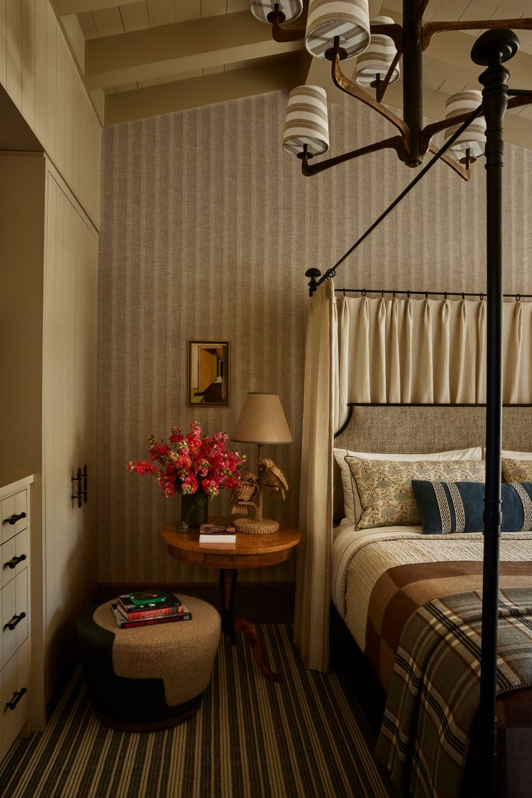 A cozy bedroom with plaid bedding on a canopy bed, beige patterned wallpaper, and striped carpet. A wooden side table holds a lamp, vase of pink flowers, books, and decor. An upholstered ottoman sits nearby; soft natural light and warm tones fill the space.