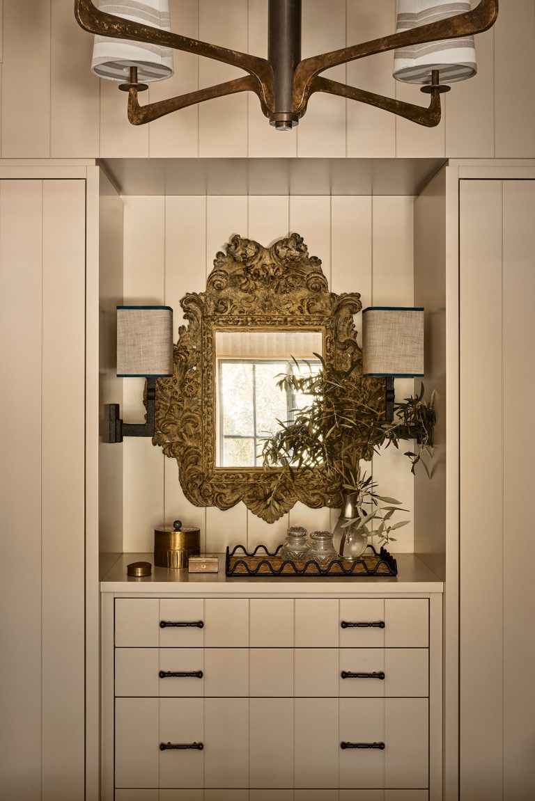 A gold ornate mirror hangs above a dresser with black handles. On the dresser are a glass vase with foliage, a decorative tray, a box, and a glass. Two wall sconces with boxy shades flank the mirror. Panel walls and a modern chandelier complete the elegant scene.