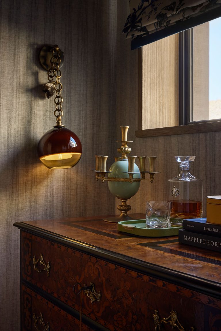 A warmly lit, elegant corner with a brown glass globe wall sconce, a gold and jade candlestick, and a decanter with amber liquid on a wooden dresser. Stacked books, a glass, and sunlight from a window add a cozy, refined ambiance to the scene.