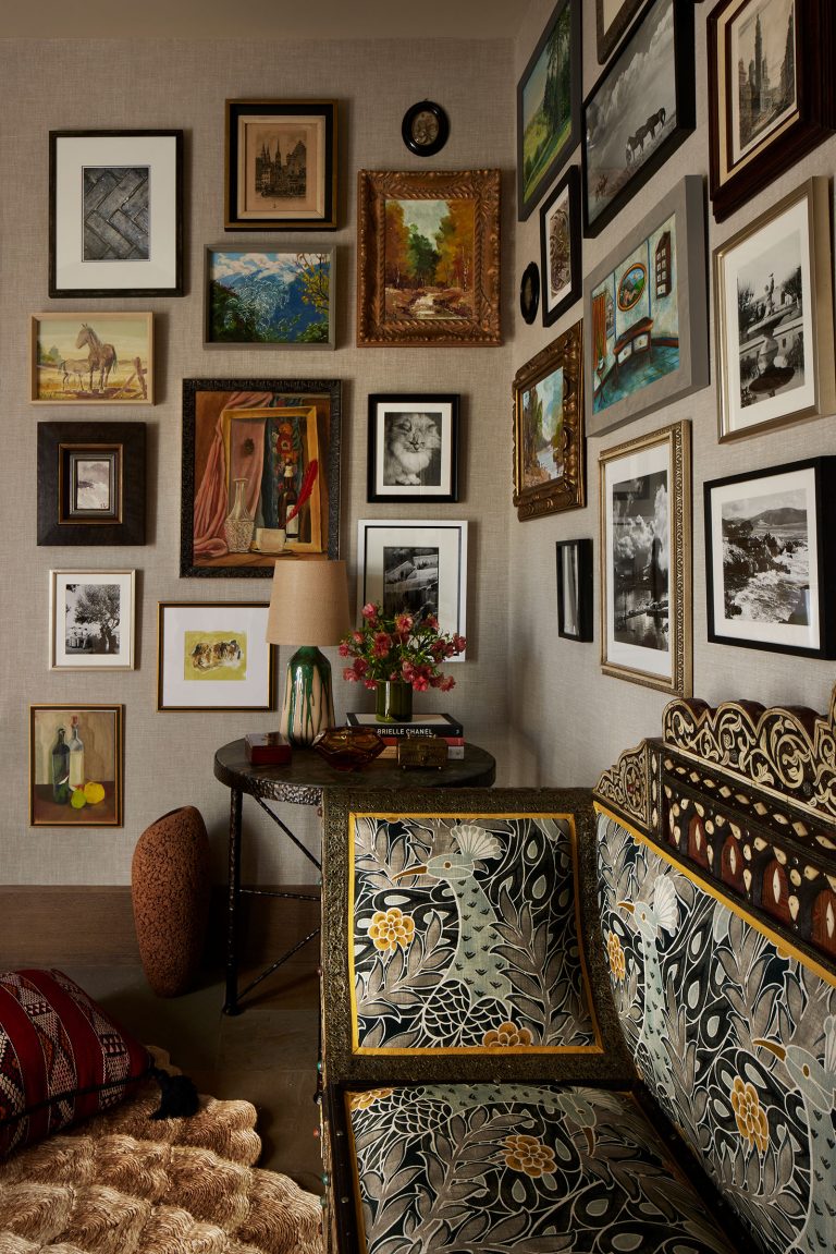 A cozy, stylish room corner features a patterned settee, a round table with a lamp and flowers, a pouf, and a wall covered in eclectic framed art and photos. Warm, earthy tones and layered textures create an inviting, artistic atmosphere.