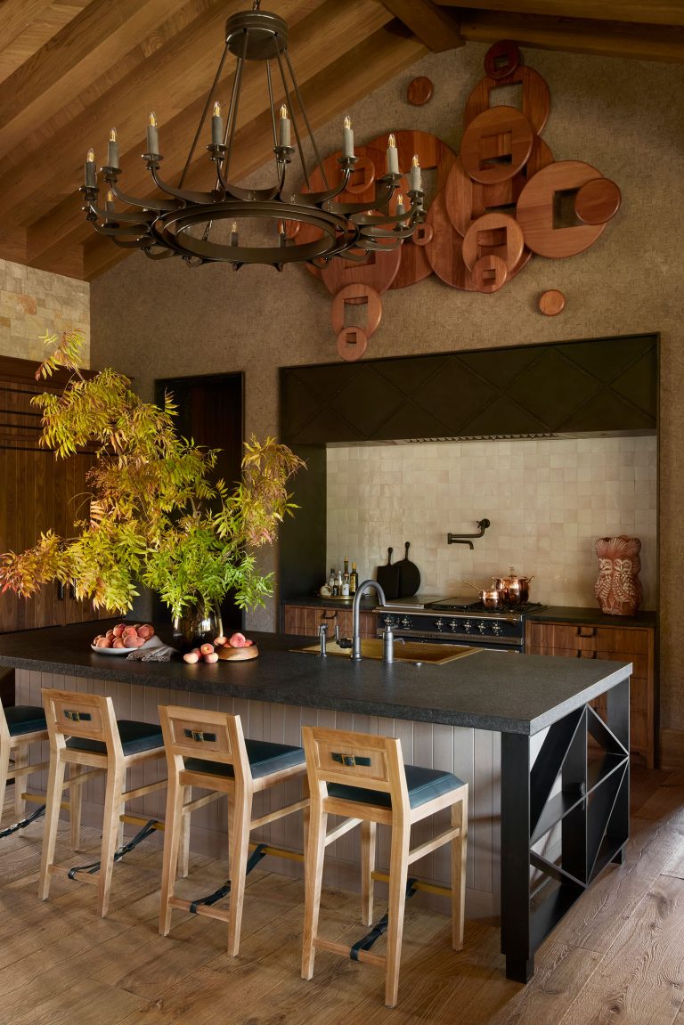 A rustic kitchen with wood beams, a large black island, four wooden chairs, and a modern chandelier. The island holds a vase with leafy branches and apples. The backsplash features neutral tiles, and the back wall displays circular wooden wall art.