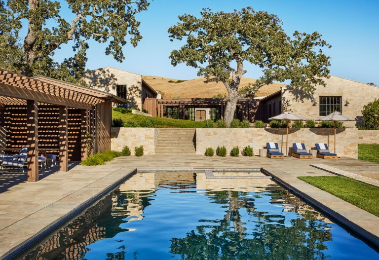 A luxurious outdoor pool reflects blue sky, bordered by stone tiles. Wooden pergola with lounge chairs on the left, sun loungers with umbrellas on the right. Stairs lead up to a rustic stone house with large trees and dry hills in the background.
