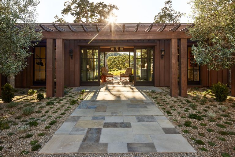 A stone path leads to a wooden pergola and glass doors of a modern house. Sunlight shines through the roof slats. Two chairs are visible inside, facing a garden with trees and open green space beyond, framed by neatly landscaped greenery.