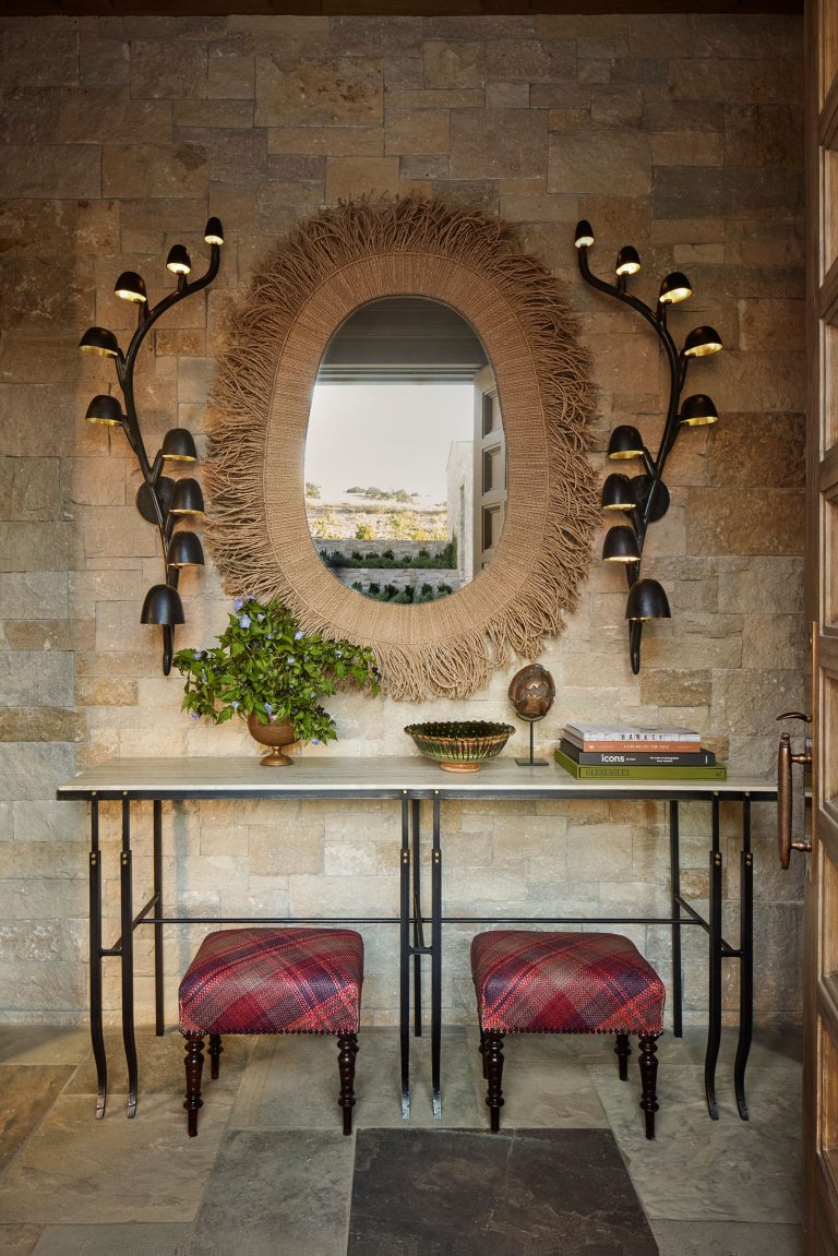 A stone wall holds an oval mirror with a woven raffia frame, flanked by black, sconce light fixtures shaped like branches. Below is a glass console table with books, a plant, and two red plaid stools with dark wooden legs on a stone tile floor.