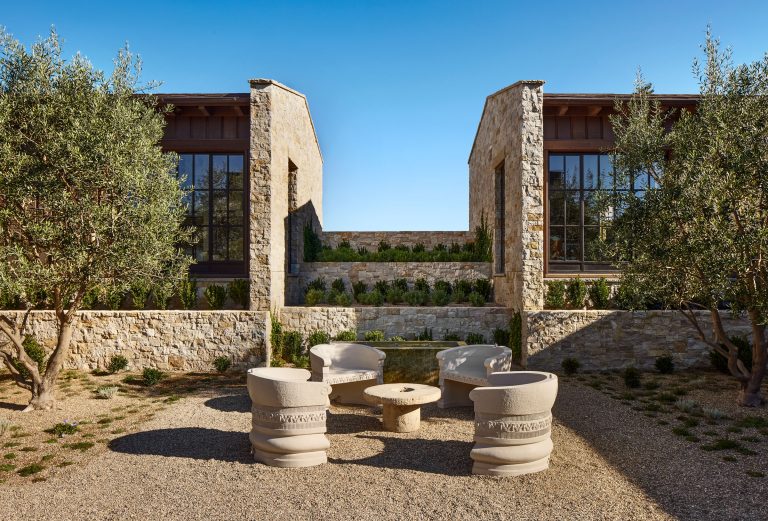 A courtyard with modern stone buildings, large windows, and two tall olive trees. In the center, four curved, light-colored chairs encircle a round stone fire pit on a gravel ground, with a tiered stone wall and manicured greenery behind. The sky is clear and blue.