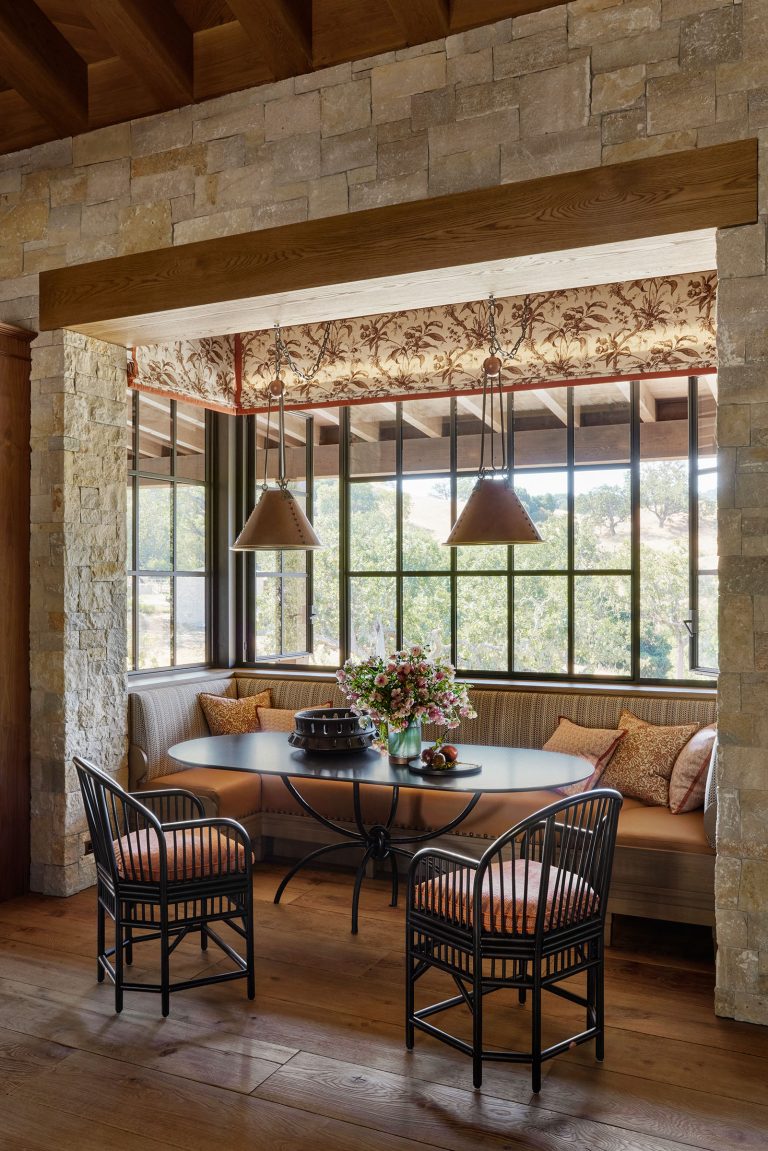 A cozy breakfast nook with a built-in cushioned bench, two black chairs, and a dark oval table topped with flowers and fruit. The nook is framed by stone walls, large windows with rustic curtains, and features two hanging pendant lamps. Warm sunlight filters in.