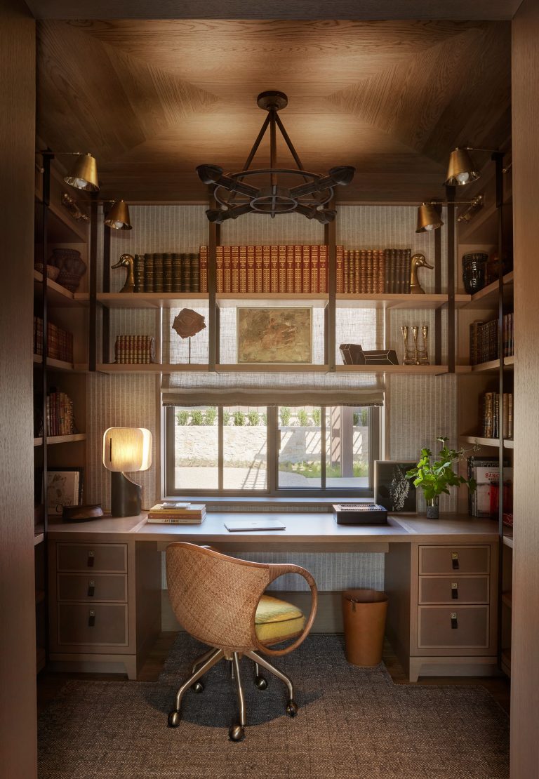 Cozy home office with a wooden desk facing a window, a rattan chair with a yellow cushion, bookshelves filled with books and decorative objects, warm lighting from sconces and a lamp, and neutral tones creating an inviting atmosphere.