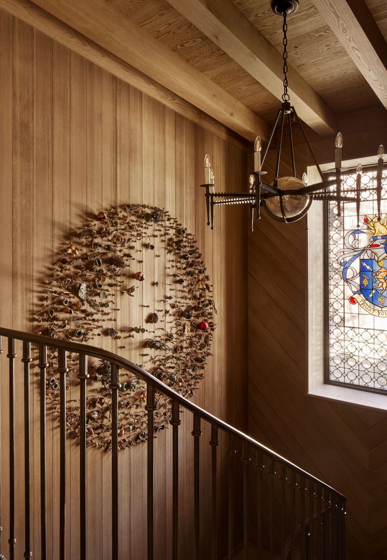 A wooden staircase with a black metal railing curves upward beside a wall covered in vertical wood panels. A textured, oval-shaped wall sculpture is mounted on the paneling. A stained glass window with a coat of arms and a metal chandelier hang above.