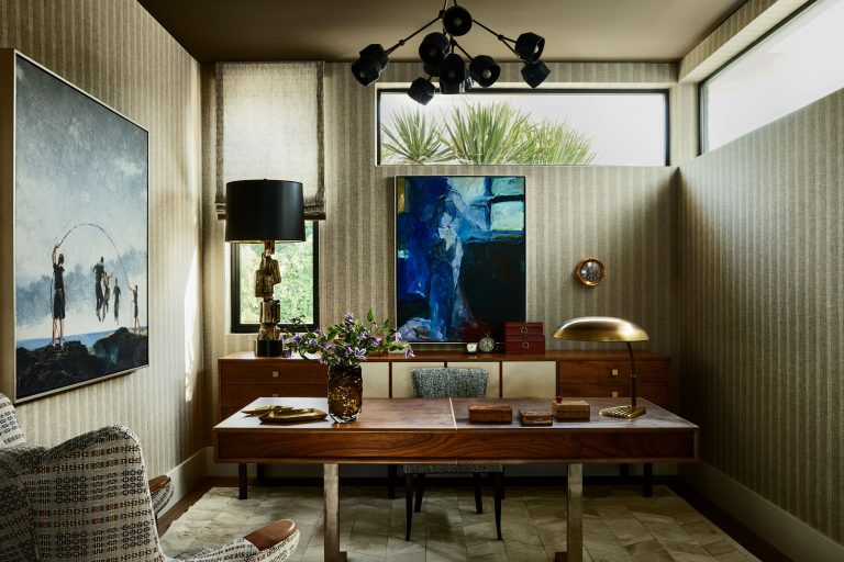 A stylish home office with a wooden desk, modern lamp, mid-century cabinet, abstract blue painting, and large window. Neutral patterned wallpaper and a mix of vintage and contemporary decor create a cozy, elegant space. A large painting and houseplants add visual interest.