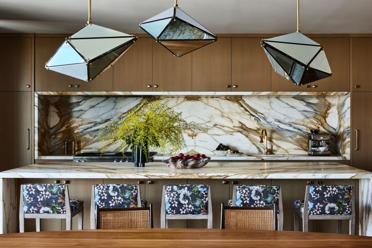 Modern kitchen with three geometric pendant lights, a marble backsplash and countertop, light wood cabinets, and four chairs with floral upholstery. A tall vase with yellow flowers and a bowl of fruit sit on the island, with a coffee machine in the background.