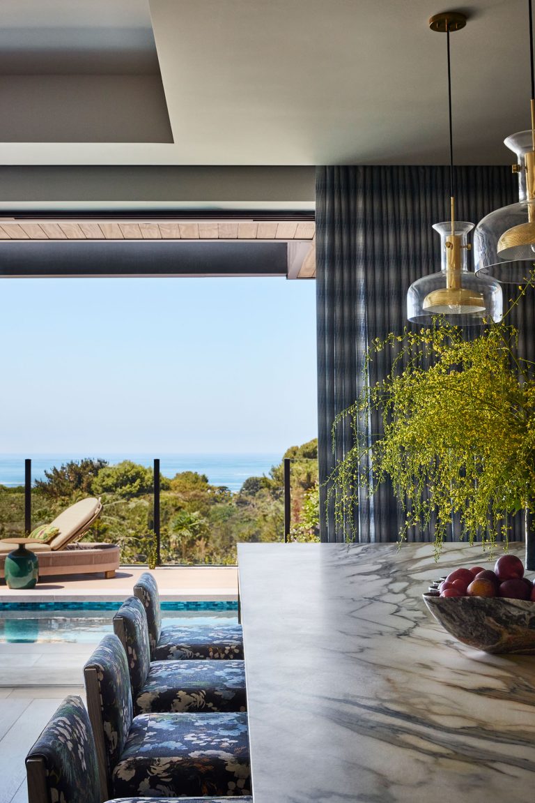 A modern kitchen with a marble island, pendant lights, floral chairs, and a bowl of fruit. Yellow flowers sit on the counter. Large glass doors open to a pool, sun lounger, trees, and an ocean view under a clear sky.
