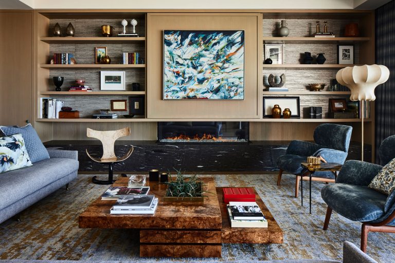 A stylish living room with a gray sofa, blue velvet chairs, a modern wood coffee table with books, a large abstract painting above a sleek fireplace, built-in shelves with decor, and a tall sculptural lamp; the room has a cozy, sophisticated feel.