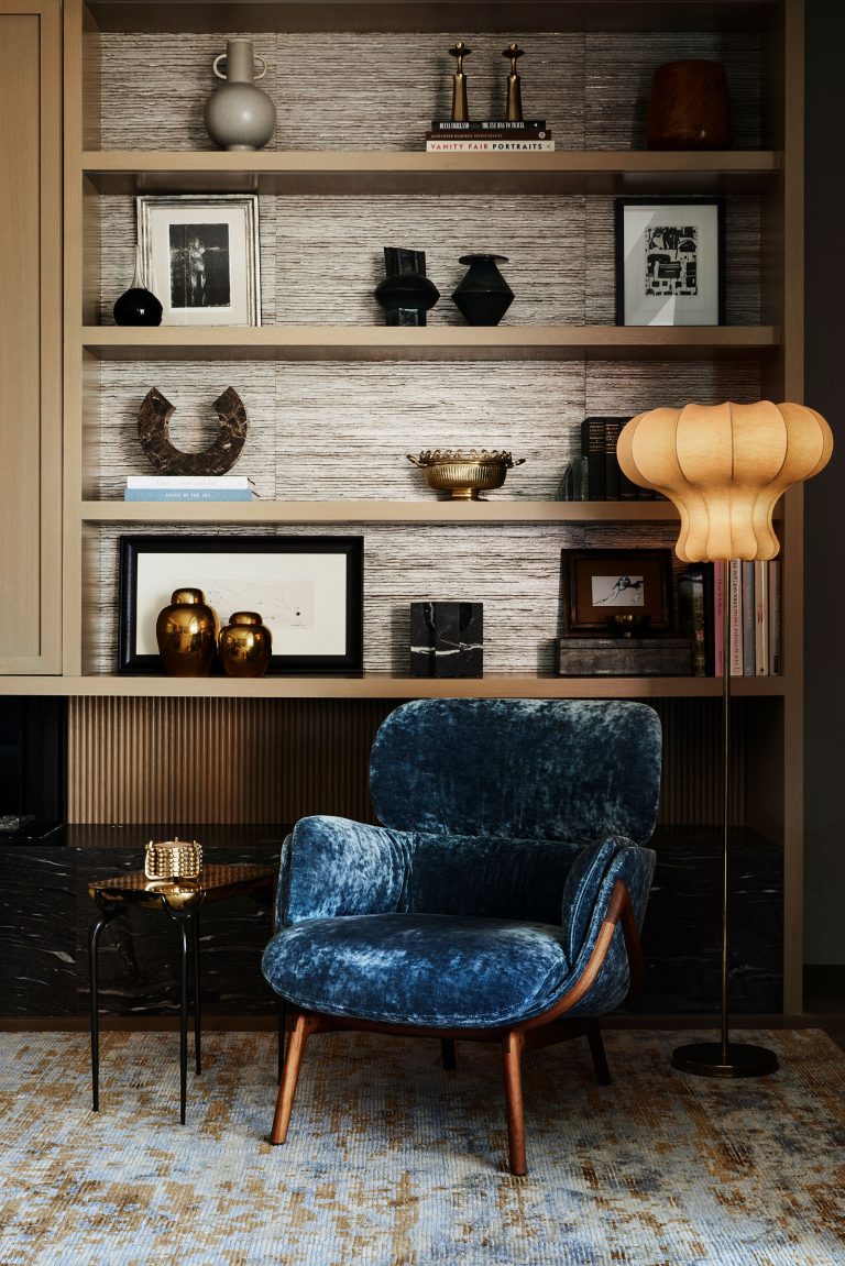 A blue velvet armchair with wooden legs sits in front of shelves filled with vases, framed photos, books, and decor. A round side table with a glass and candle is beside the chair. A tall, sculptural floor lamp glows warmly beside the chair.