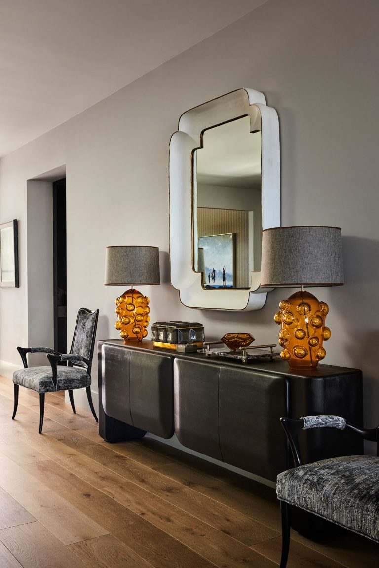 A modern hallway with light wood floors features a black console table topped with two amber glass bubble lamps, a tray, and a decorative bowl. Above is a large, sculptural wall mirror. Two black armchairs with patterned upholstery flank the table.