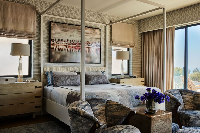 A modern bedroom with a white four-poster bed, gray bedding, and two wooden nightstands holding lamps. Large windows with beige shades flank the bed. Two patterned armchairs face a small table with a vase of purple flowers. A large art piece hangs above the bed.