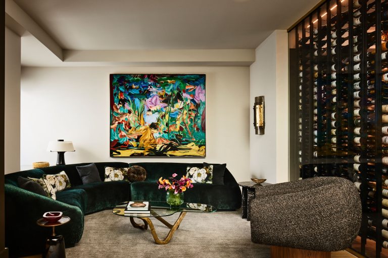 Modern living room with a dark green curved velvet sofa, glass coffee table, abstract floral paintings, and a textured armchair. A wine wall is visible on the right, and a vase with colorful flowers is on the table. The room has neutral walls and soft lighting.