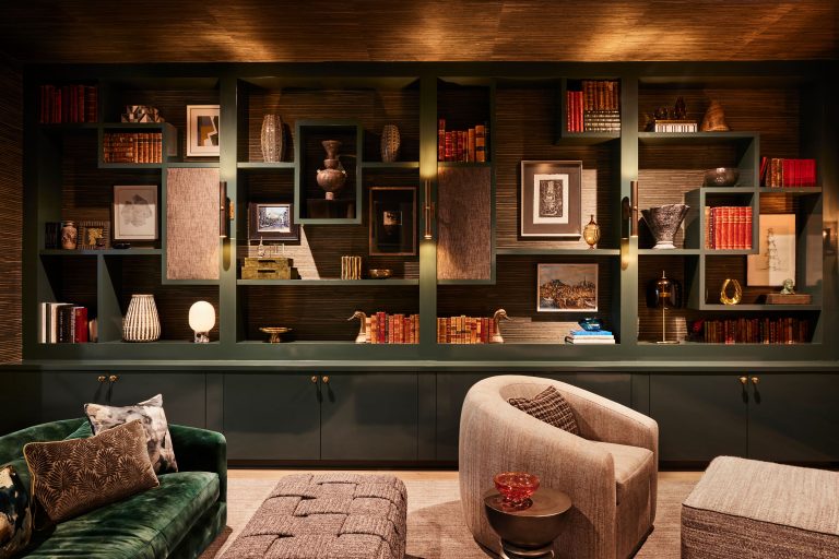 A cozy library with green built-in bookshelves filled with books, framed art, vases, and decorative objects. Plush seating includes a green velvet sofa, a gray armchair, and an upholstered bench, all on a neutral rug. Warm lighting creates a welcoming atmosphere.