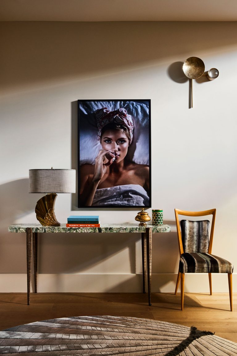 A modern interior features a marble console table with books, a lamp, and a mug. Above it hangs a framed photo of a woman in bed. To the right, a velvet-striped chair sits under a sculptural gold wall sconce. The floor has a geometric-patterned rug.