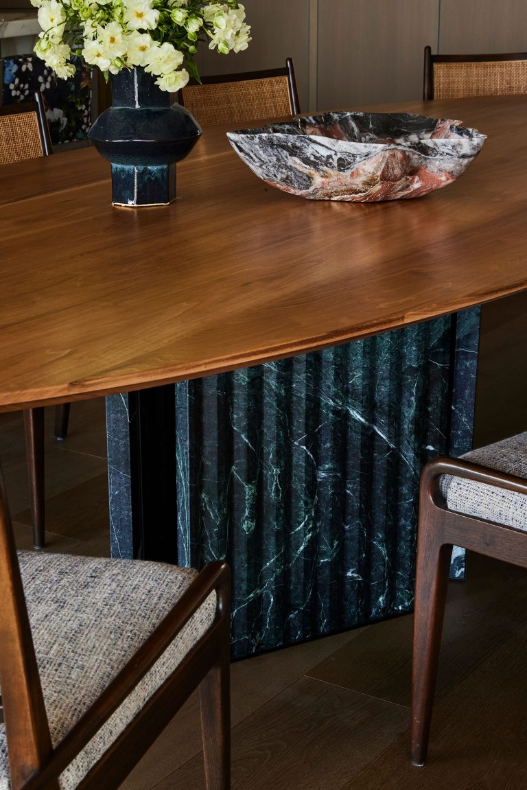 A wooden dining table with a dark green marble base stands in a modern room. On the table are a black vase of white flowers and a large marbled bowl. Wooden chairs with light upholstered seats surround the table.