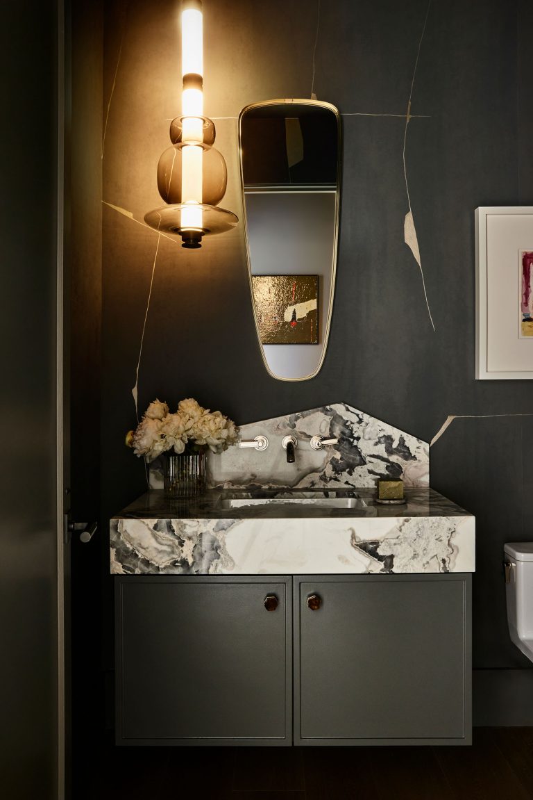A modern bathroom with dark marble-patterned sink, gray vanity cabinet, wall-mounted faucet, oval mirror, vertical light fixture, white flowers in a vase, soap dish, and dark walls with light veining. Framed art hangs on the right wall above the toilet.
