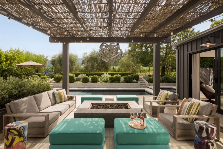 A modern outdoor patio with cushioned sofas and turquoise ottomans around a square fire pit, beneath a wooden pergola. String lights hang overhead, and lush greenery, a pool, and a shaded seating area are visible in the background.