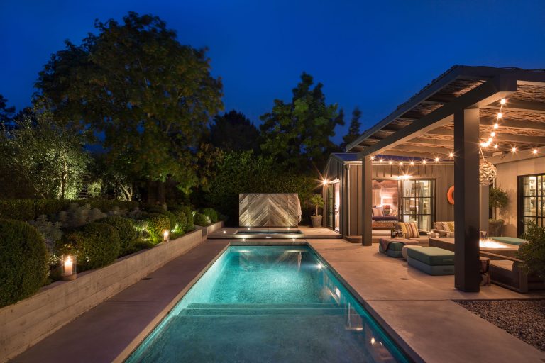 Modern backyard at dusk featuring a narrow illuminated swimming pool, elegant landscaping with trimmed bushes, and a covered patio area. Patio has cozy seating and string lights, while the pool reflects surrounding greenery and architecture.