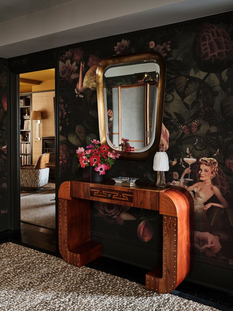 An art deco console table with a vase of pink flowers, a small lamp, and books sits under a large, vintage gold-framed mirror. The wall behind features dark, whimsical wallpaper with floral and vintage female figure motifs. A woven rug covers the floor.