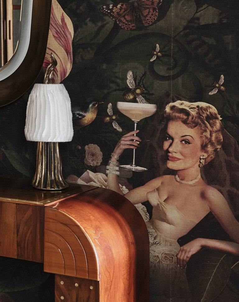 A vintage-style wallpaper depicts a glamorous woman in a strapless dress holding a cocktail. She is surrounded by butterflies and birds. In front is a shiny wooden console table and a gold lamp with a white pleated shade. A mirror edge is visible on the left.