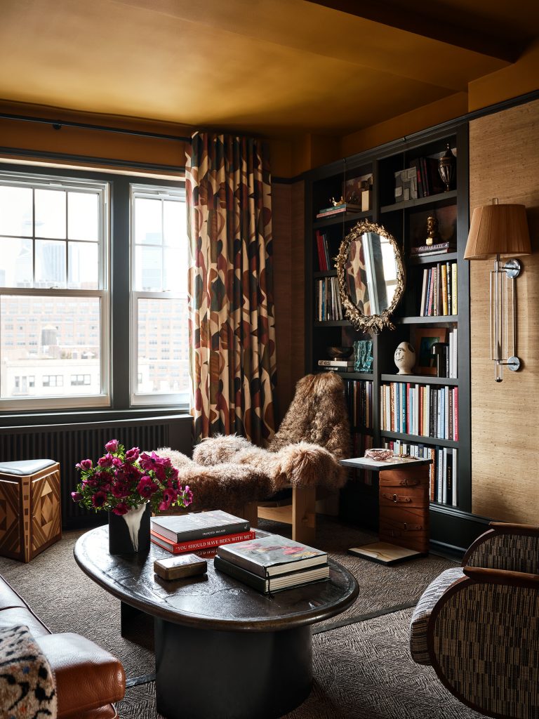 A cozy, stylish living room with large windows and city views. There’s a furry chaise lounge by a black bookshelf with books, art, and a round mirror. A coffee table with books and a flower vase sits on a patterned rug. Warm, brown tones and patterned curtains complete the space.