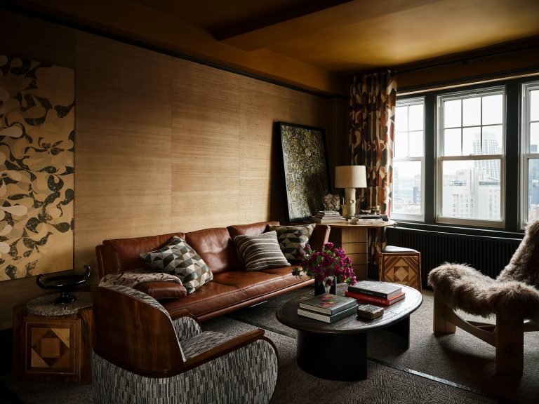 A cozy living room with a brown leather sofa, patterned pillows, wooden armchairs, a round black coffee table with books and flowers, textured wallpaper, abstract artwork, a city view through large windows, and warm, earthy tones throughout.