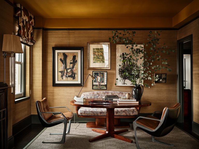 A cozy, stylish home office with brown grasscloth walls, a wooden round table, cushioned bench, two leather chairs, framed art on the wall, a black vase with branches, books, a lamp, and sunlight from a window with patterned curtains.