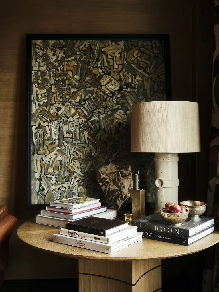 A round wooden table holds stacked art books, a cream ceramic lamp with a beige shade, a small abstract sculpture, a brass bowl, a candle, and a plate of figs. Behind, a large framed abstract painting with earthy, textured brushstrokes leans on the wall.