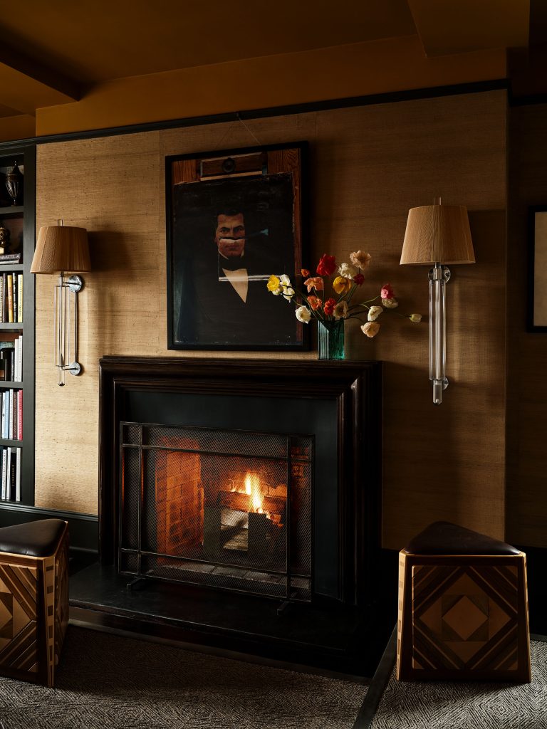 A cozy living room features a lit fireplace set in a black mantel, flanked by bookshelves and sconces with beige lampshades. Above the mantel hangs a dark portrait. A vase with colorful flowers sits beside the fire, and two geometric stools are nearby.