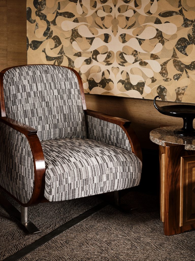 A vintage armchair with curved wooden arms and patterned black-and-white upholstery sits beside a small, round, marble-top table. Behind it is a beige and black abstract wall art with organic, swirling shapes. The setting has a warm, retro feel.