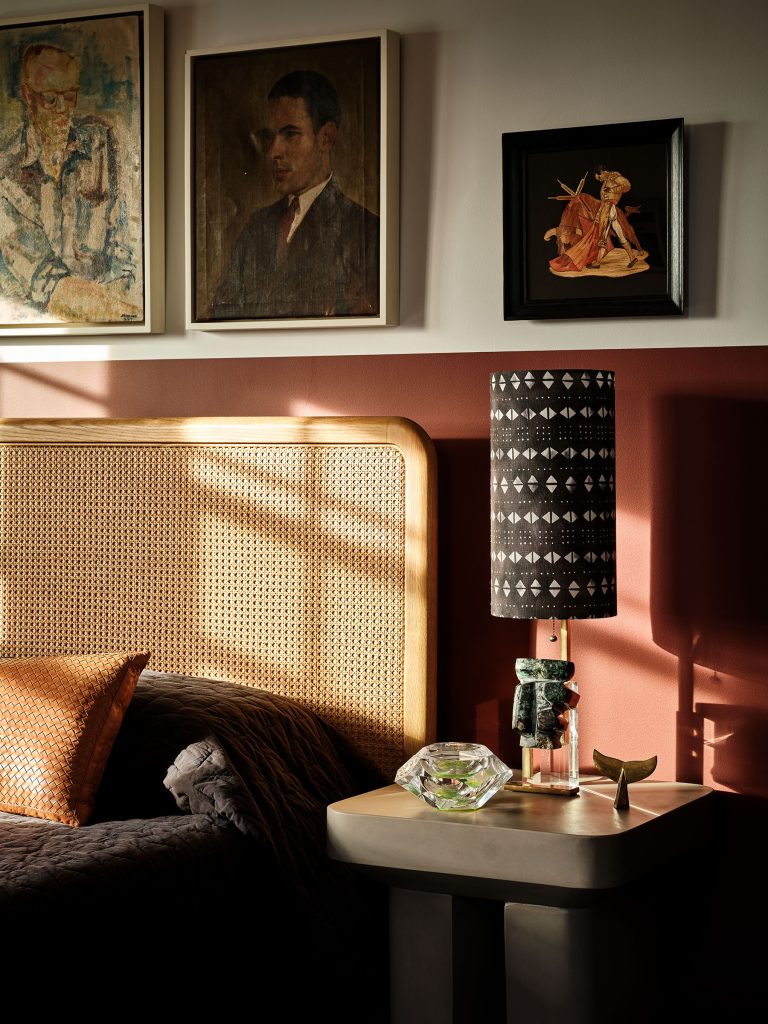 A stylish bedroom corner with a woven rattan headboard, burnt orange accent wall, patterned lamp, geometric side table, glass decor, and framed art above—two portraits and a small painting of a woman in traditional dress. Sunlight casts shadows across the scene.