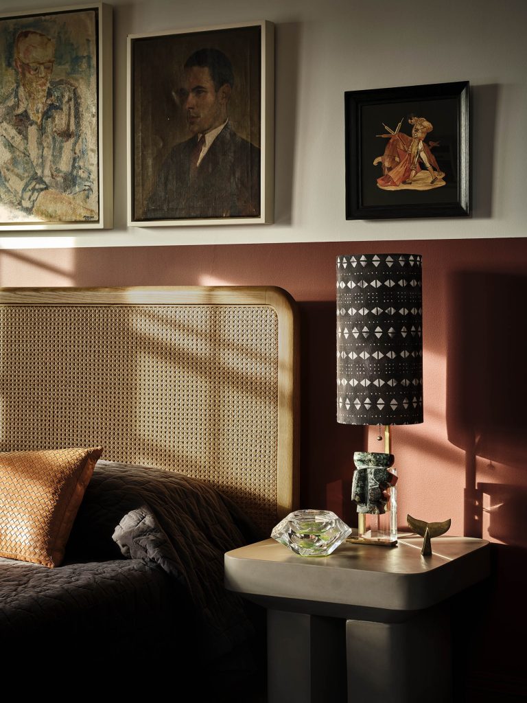 A cozy bedroom corner features a rattan headboard, dark bedding, and an orange textured pillow. On a modern nightstand sits a glass lamp with a patterned black-and-white shade, a glass ornament, and a brass bird. Three framed artworks hang above. Sunlight casts shadows.
