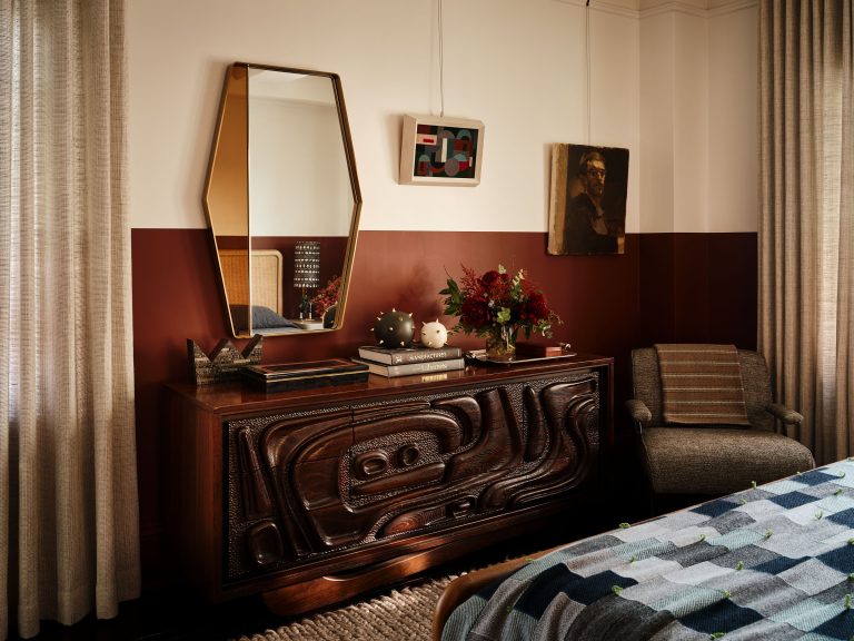 A stylish bedroom with a dark wood, carved dresser topped with books, vases, and flowers. Above it are an angular mirror, abstract art, and a portrait. To the right is a gray upholstered chair; beige curtains and a bed with a patterned blanket are partially visible.