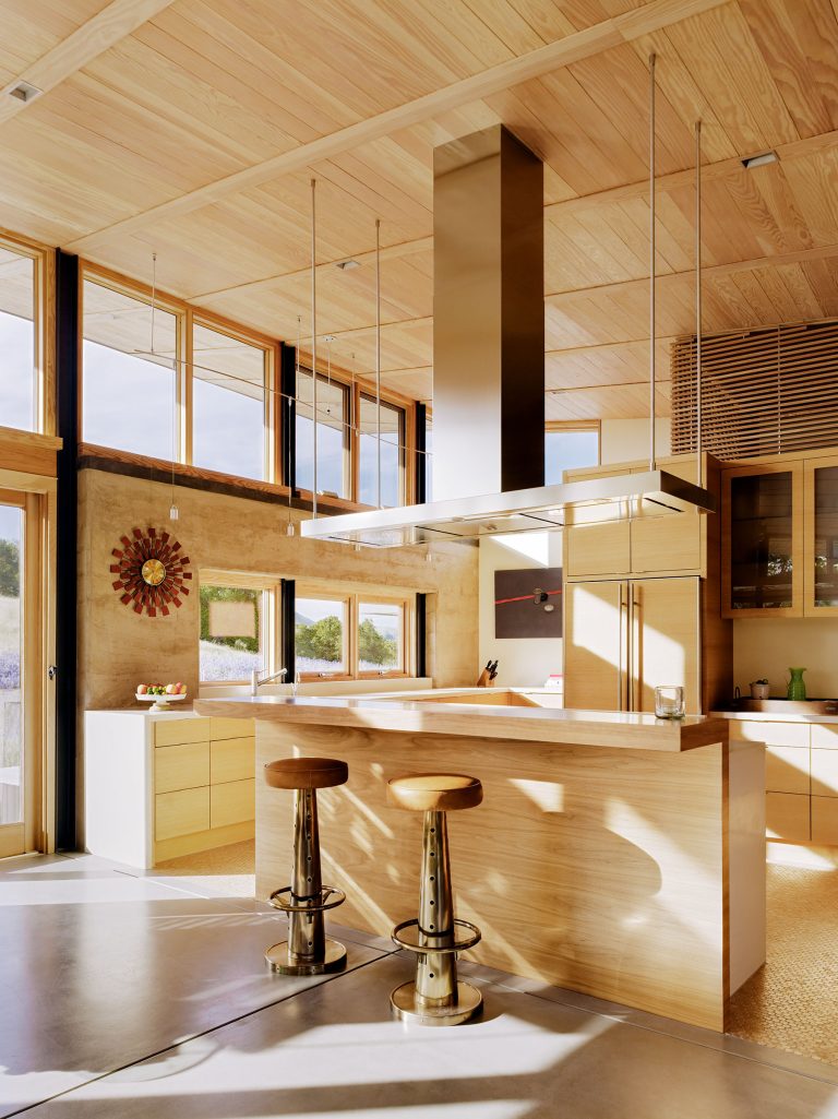 A modern kitchen with light wood walls, ceiling, and cabinetry. An island with two gold and wood bar stools sits under a large stainless steel range hood. Large windows let in natural light; a decorative wall clock and open shelves add accents.