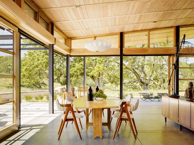 A modern dining room with large floor-to-ceiling windows, light wood ceiling and furniture, and views of green trees outside. The space features a rectangular wooden table with six chairs, a pendant lamp above, and natural sunlight streaming in.