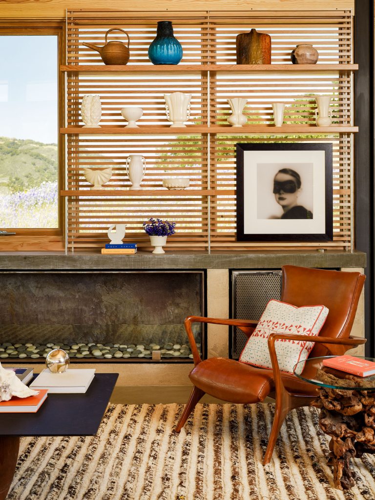 A cozy living room with a tan leather armchair, striped rug, and a modern fireplace. Wooden shelves display white vases, pottery, and a blue vase. A framed black-and-white portrait sits on the mantel, with a scenic view visible through the window.