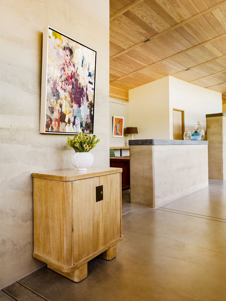 A modern, minimalist interior with light wood accents. A colorful abstract painting hangs above a small wooden cabinet topped with a white vase of yellow flowers. In the background, a stone reception desk and a hallway are visible. The space is bright and airy.
