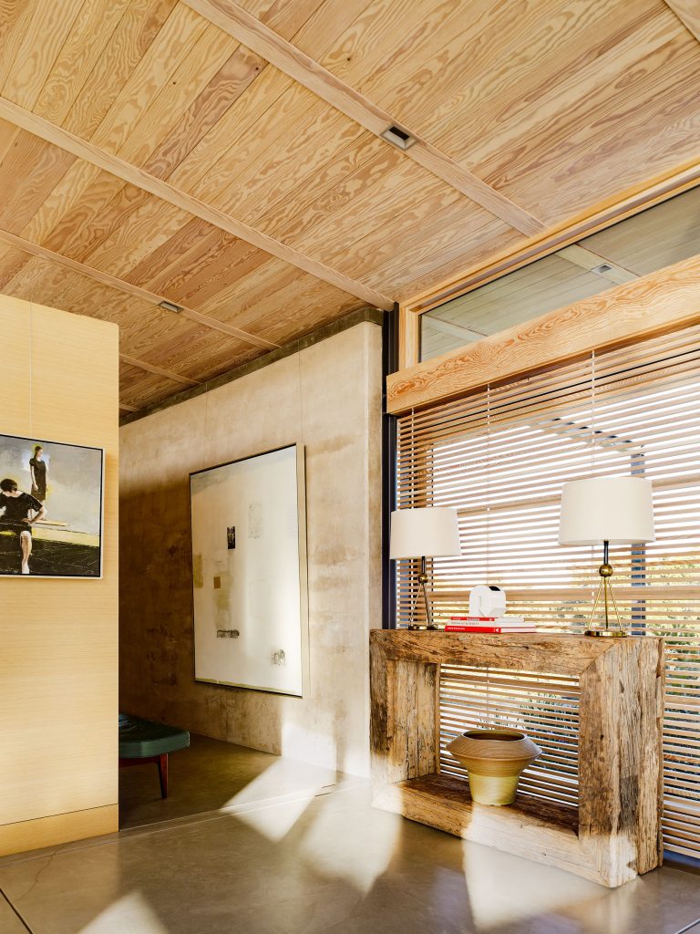 A sunlit modern interior features a rustic wooden console table with two lamps and decor, next to large windows with wooden blinds. Art hangs on textured beige walls, and a wooden ceiling enhances the warm, natural atmosphere.