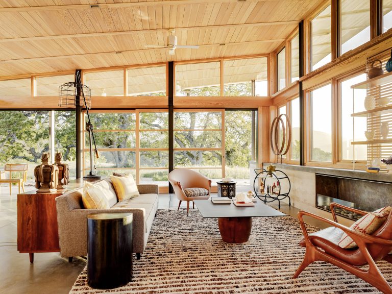 A modern living room with large glass windows, wood ceiling, and neutral-toned furniture. The space features a gray sofa, accent chairs, a patterned rug, and artful decor. Natural light floods the room, highlighting trees and nature visible outside.