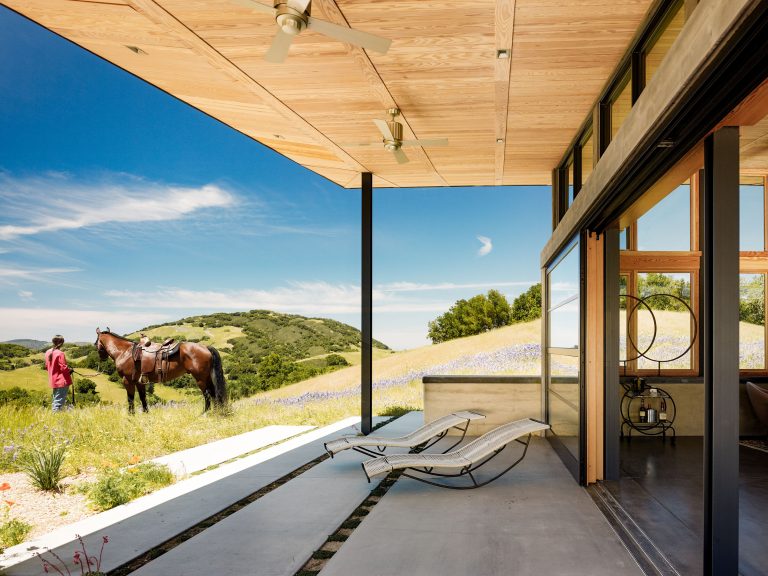Modern home patio with two lounge chairs overlooks grassy hills under a blue sky. A person stands beside a saddled horse among wildflowers. Ceiling fans hang above the patio, and large glass doors connect the outdoor and indoor living spaces.
