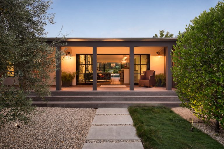 A modern single-story house features a covered porch with three columns, warm exterior lighting, large glass doors, and windows. The landscaped front yard has gravel, stepping stones, grass, and lush green bushes framing the entrance.